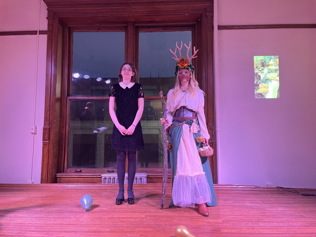 Two participants stand on stage during a special effects makeup competition. One is dressed in a black dress with lace sleeves, while the other is adorned in a fantasy costume with antlers and a floral crown, holding a walking stick and a basket.