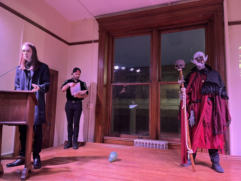 A performance space showcasing a special effects makeup competition, featuring a speaker at a podium, a judge holding papers, and a contestant in a dramatic costume with skeletal elements.