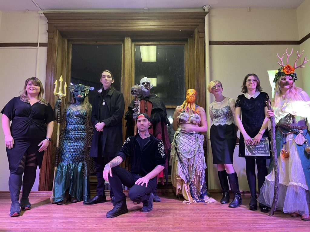 Group of special effects makeup artists posing together in front of a wooden backdrop, showcasing their creative costumes inspired by sea creatures, forest guardians, and fantasy elements.