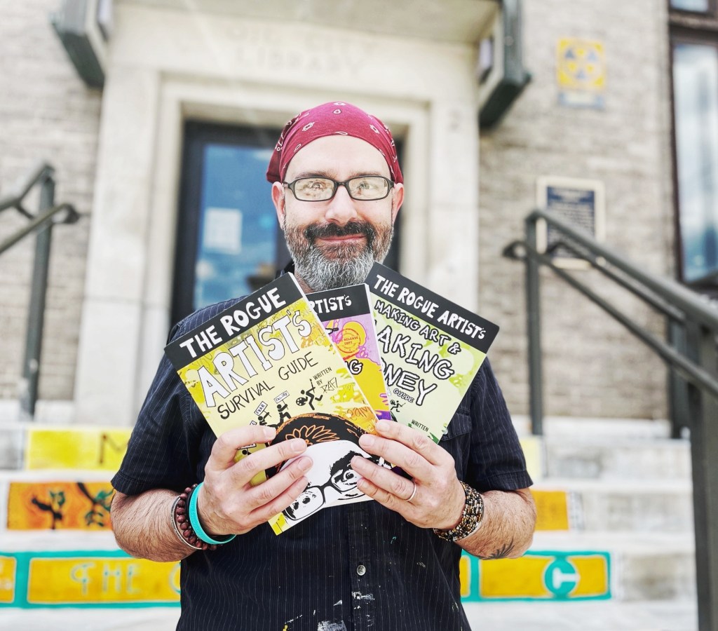 A man with glasses and a bandana stands outside a library holding three books titled 'The Rogue Artist's Survival Guide', 'Making Art', and 'Making Money'.