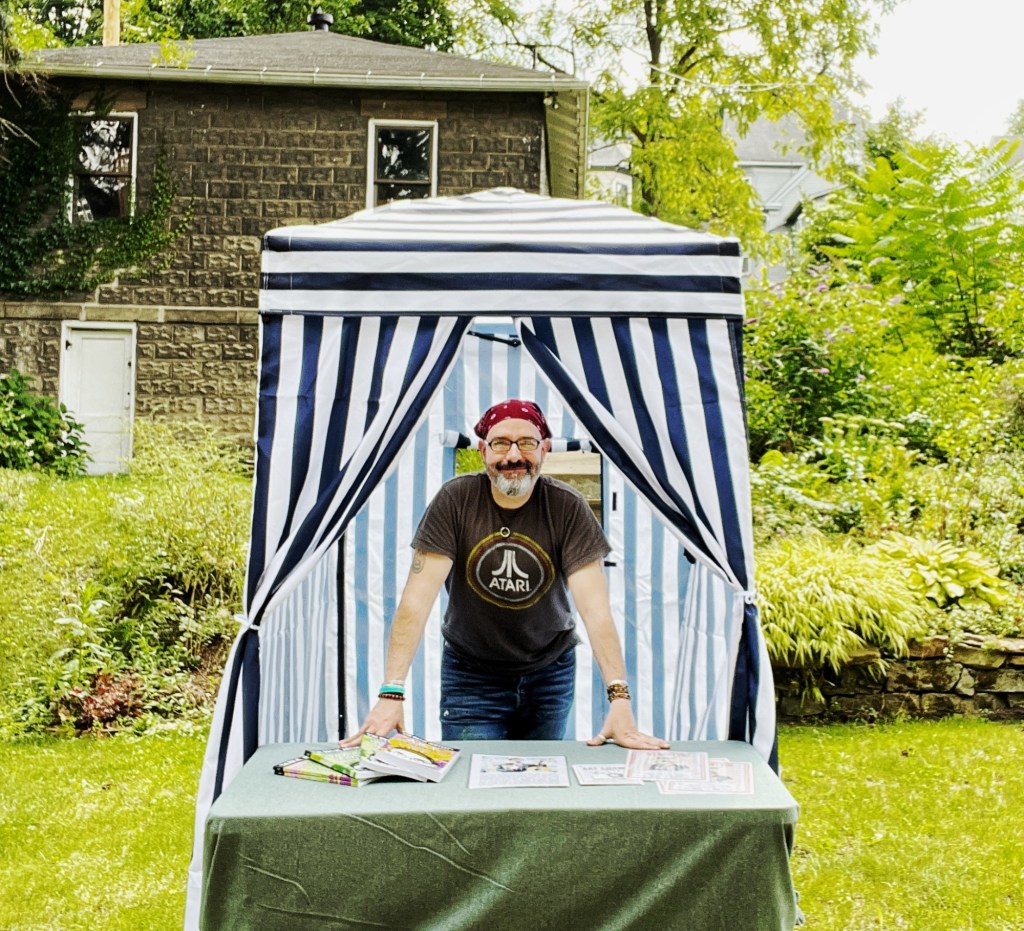 A man in a red bandana and an Atari t-shirt leans on a table at a striped booth in a green outdoor setting, promoting his artistic books.