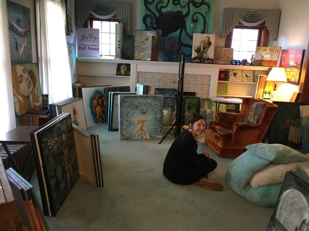 A woman sitting on the floor in an art-filled living room, surrounded by various paintings and artwork, with a cozy armchair and decorative items in the background.