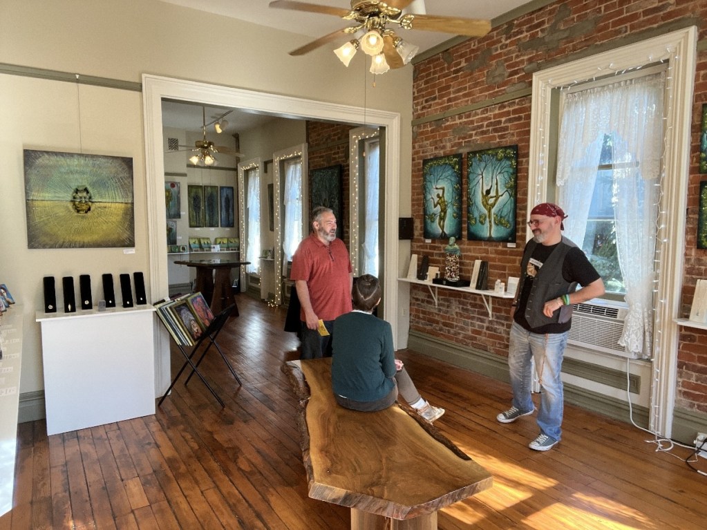 Three people interacting inside an art gallery with numerous paintings displayed on the walls and a wooden table in the center.