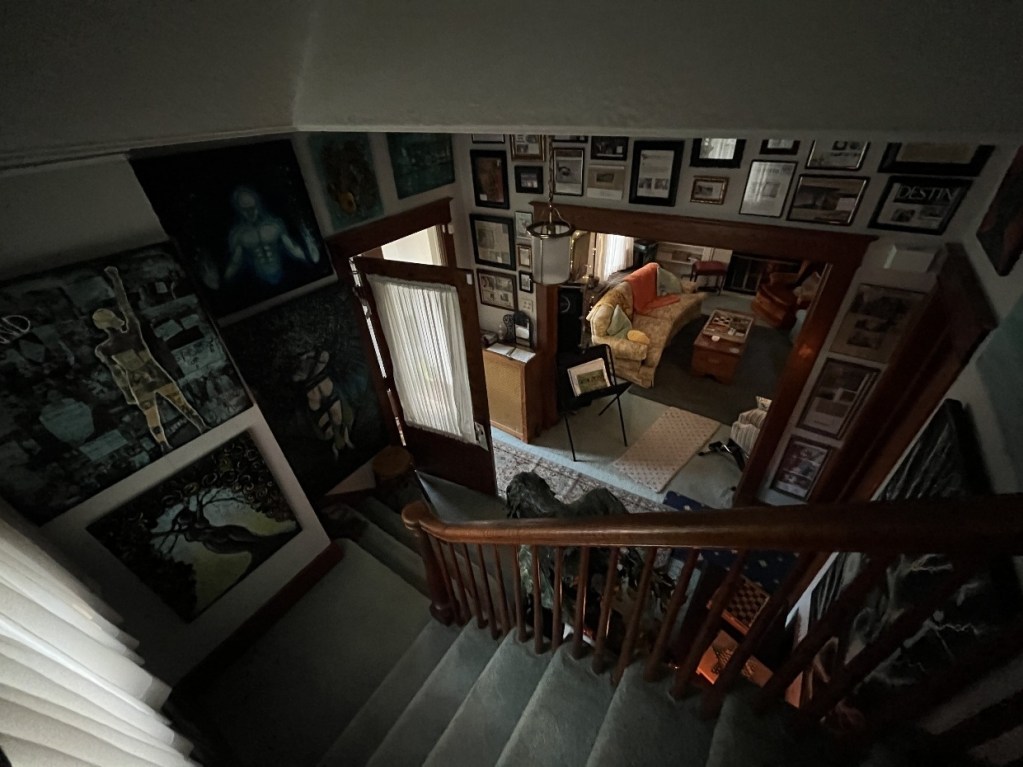 An overhead view of a staircase leading down to a cozy living room filled with framed artwork and photographs, showcasing a creative and artistic environment.