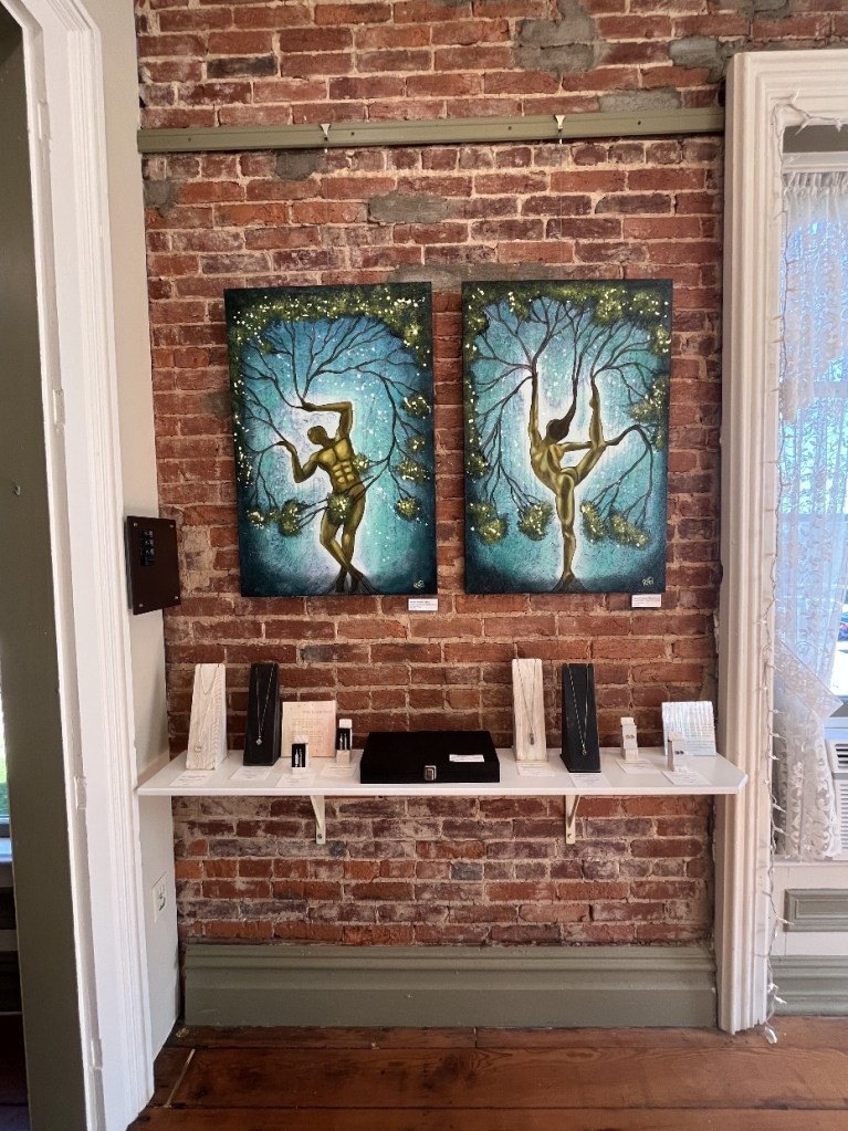 Artistic display featuring two paintings of dancers intertwined with tree branches, set against a brick wall. A shelf below holds various artistic items and jewelry.