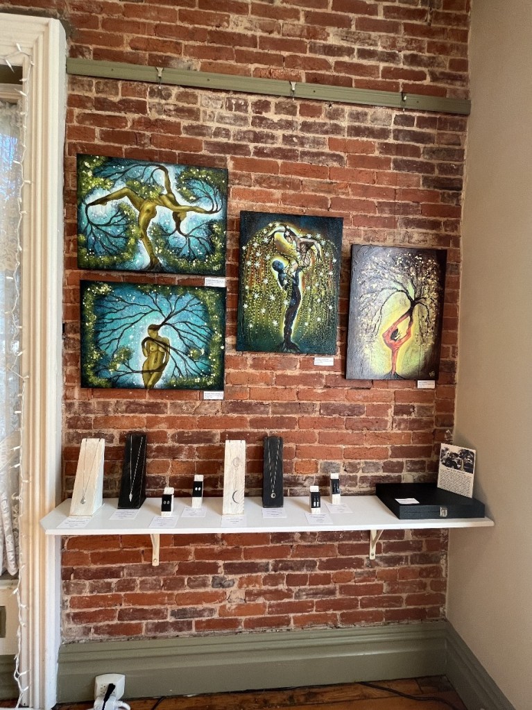 Art display featuring four colorful paintings on a brick wall, with a shelf below showcasing various jewelry pieces and information cards.