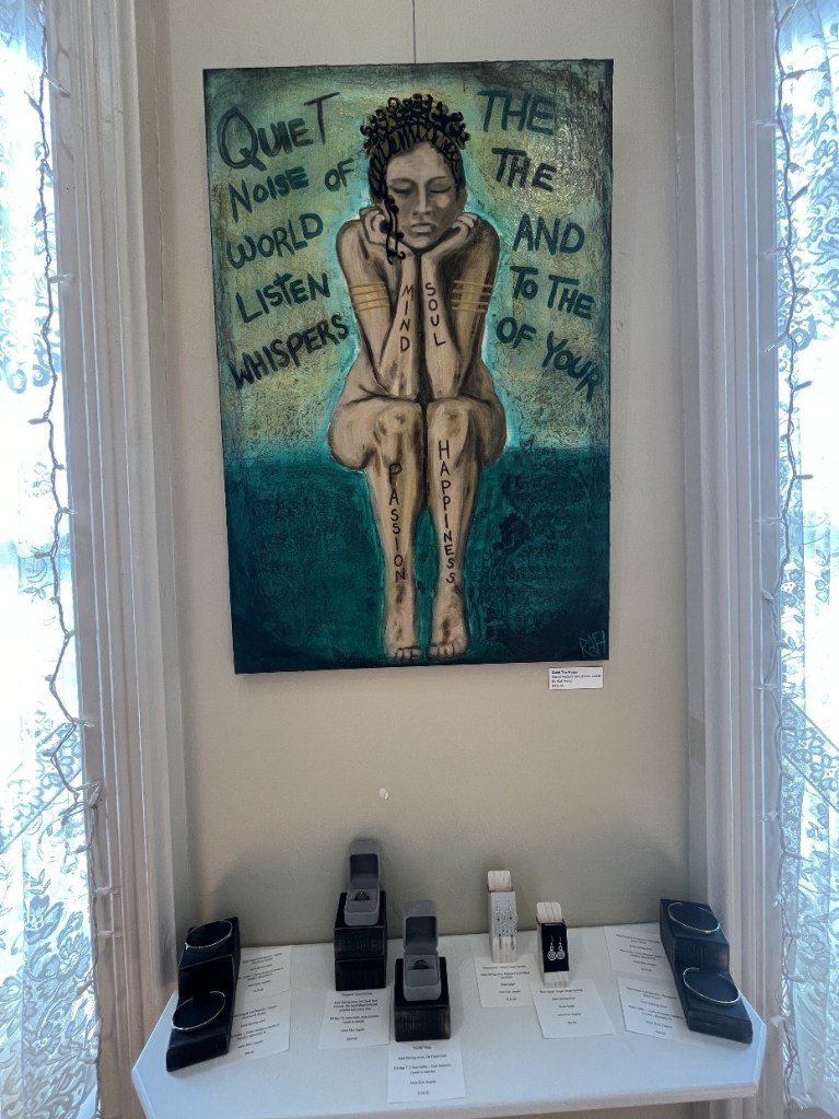 A painting of a seated woman with intricate hair, surrounded by the words 'Quiet the noise of the world, listen to the whispers of your mind, soul, passion, and happiness.' Below the painting, a display of handcrafted jewelry pieces is arranged on a table.