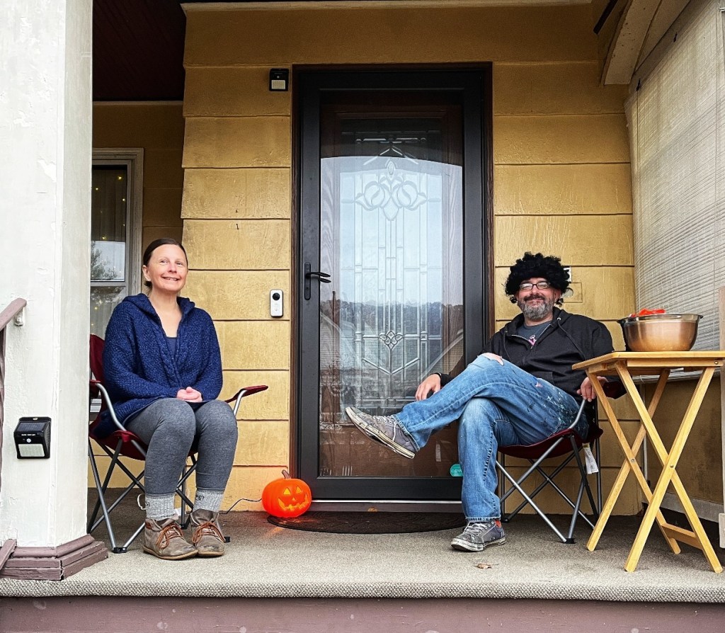 Two people sitting on a porch, one wearing a curly wig, smiling beside a carved pumpkin and a table with treats.