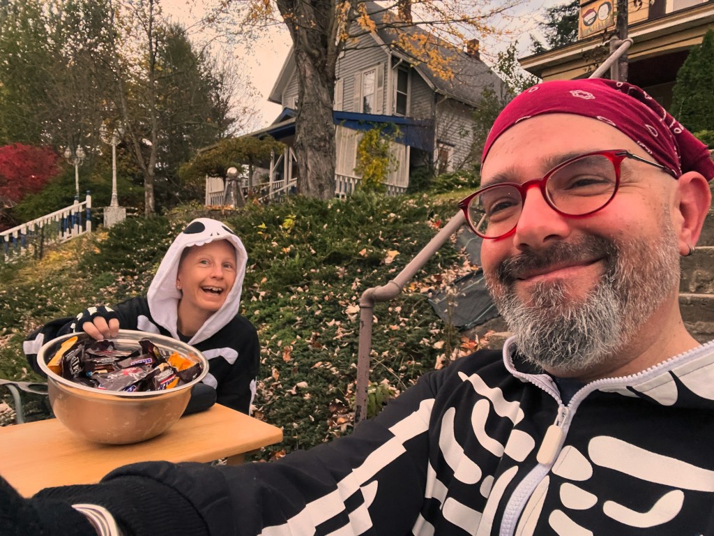 A cheerful moment captured on Halloween, featuring a person and a child in skeleton costumes, sitting outside with a bowl of candy. The background showcases autumn foliage and a house.