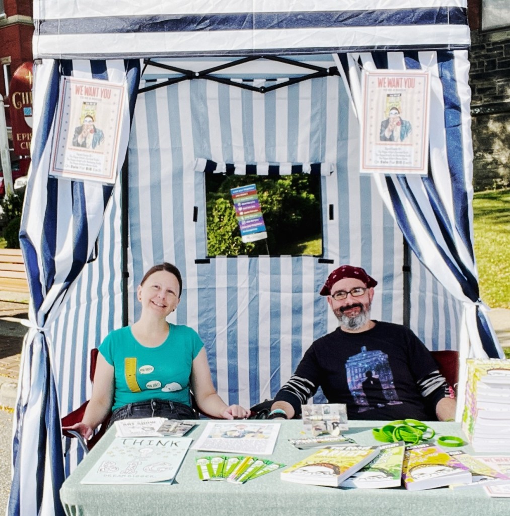 Two artists seated at a booth with a striped canopy, displaying various art materials and promotional items on the table.