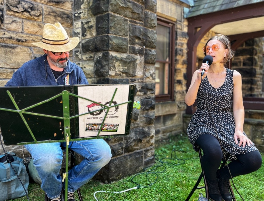 A musician playing guitar while a woman sings into a microphone in an outdoor setting, with a stone wall in the background.