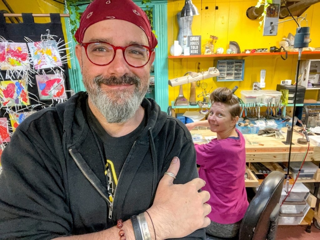 A smiling man in a black hoodie poses with crossed arms in front of a woman working at a craft table, both surrounded by colorful artwork and art supplies in a vibrant studio.