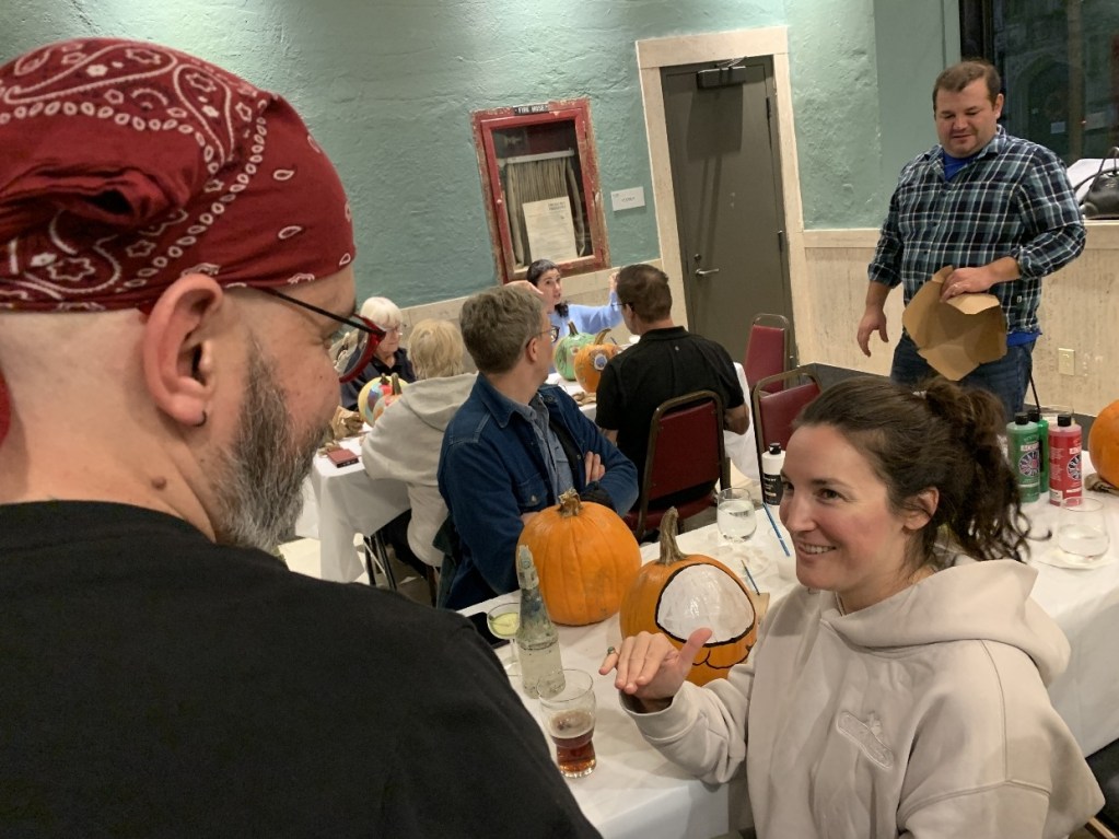 Group of people engaged in a pumpkin painting party, with one person in conversation and pumpkins on the table.