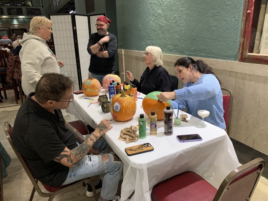 A group of people engaged in a pumpkin painting activity, surrounded by brushes, paints, and completed pumpkin designs on a table.
