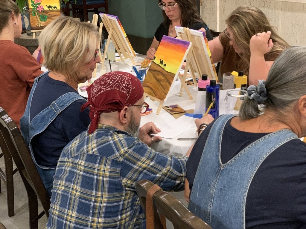 Participants engaged in a Paint and Sip event, focused on creating art on mini canvases, with various art supplies visible on tables.