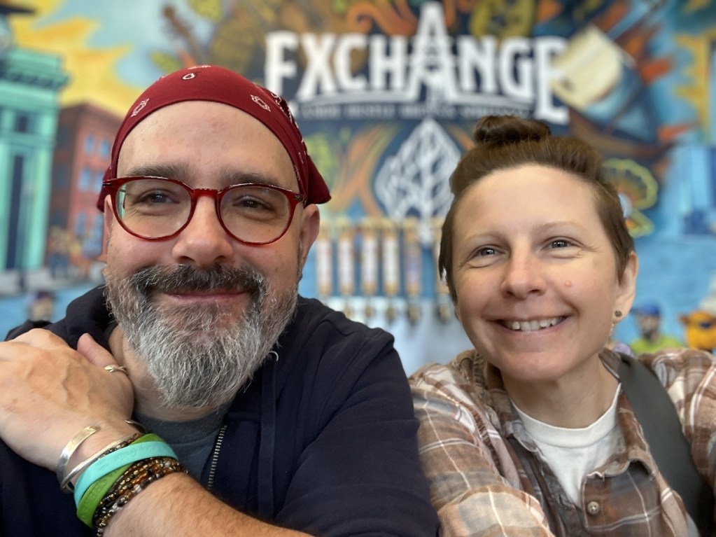 A smiling man with a beard and glasses, wearing a red bandana, sits next to a woman with short hair and a plaid shirt. They are in a colorful venue with 'EXCHANGE' written behind them.