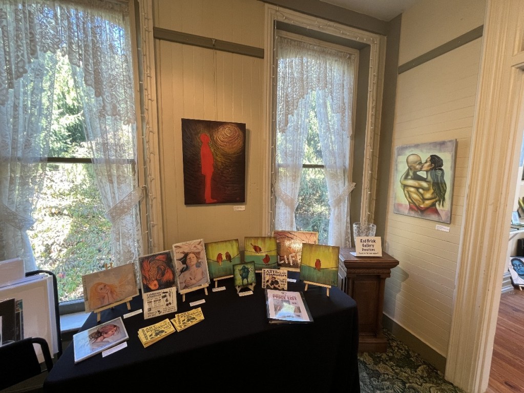 Gallery exhibition featuring various art pieces on display, including paintings and jewelry, with lace curtains and natural light coming through windows.
