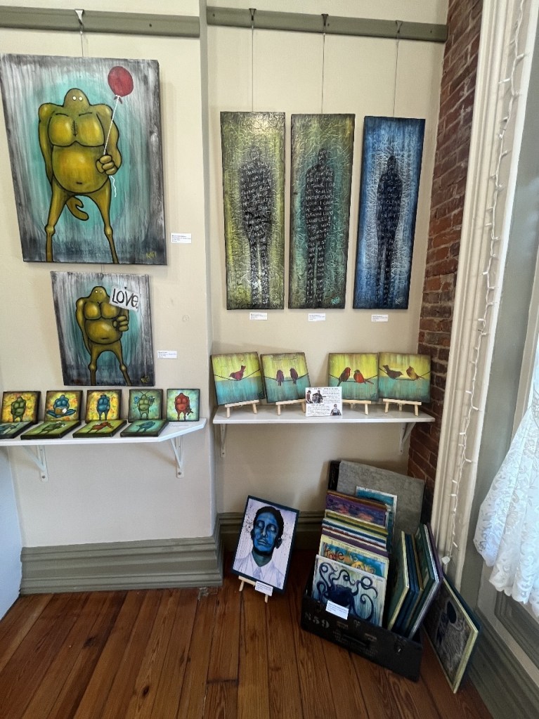 Gallery corner featuring colorful artwork, including a large painting of a character holding a balloon, abstract paintings with silhouettes, and smaller framed pieces displayed on shelves against a brick wall.