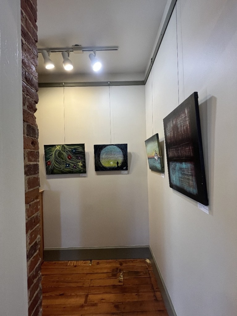 Interior view of an art gallery showcasing several framed paintings on the walls, with exposed red brick and wooden flooring.