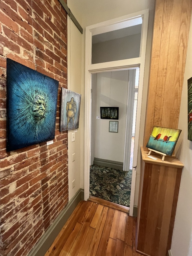 An interior view of an art gallery featuring brick walls and wooden flooring. Several paintings are hung on the walls, showcasing various artistic styles and colors. A small wooden display stand holds a painting on the right, while a doorway leads to another room in the background.