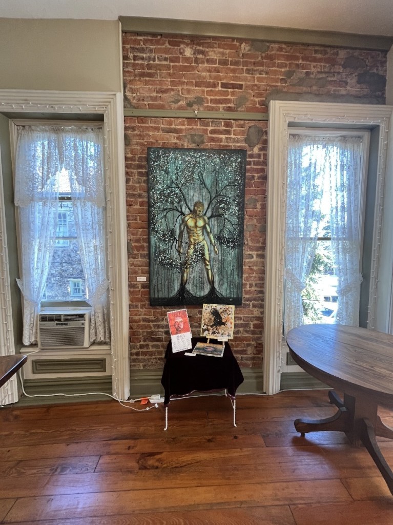 An exhibit space featuring a large art piece of a figure against a brick wall, accompanied by small displays on a table, with lace curtains in the windows letting in natural light.