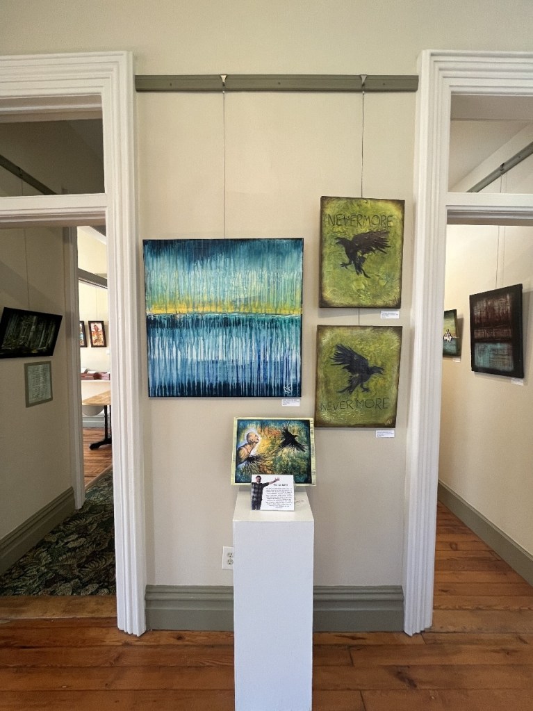 An art gallery exhibition featuring multiple paintings hanging on the walls and a pedestal with an artwork and a printed description in the foreground. The walls are painted light colors with wood flooring and decorative elements, creating an inviting atmosphere.