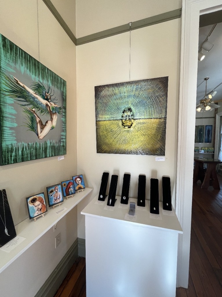 An art gallery showcasing vibrant paintings, including a piece of a dancer with feathers and other artwork, alongside displays of handcrafted jewelry and small framed portraits on a shelf.