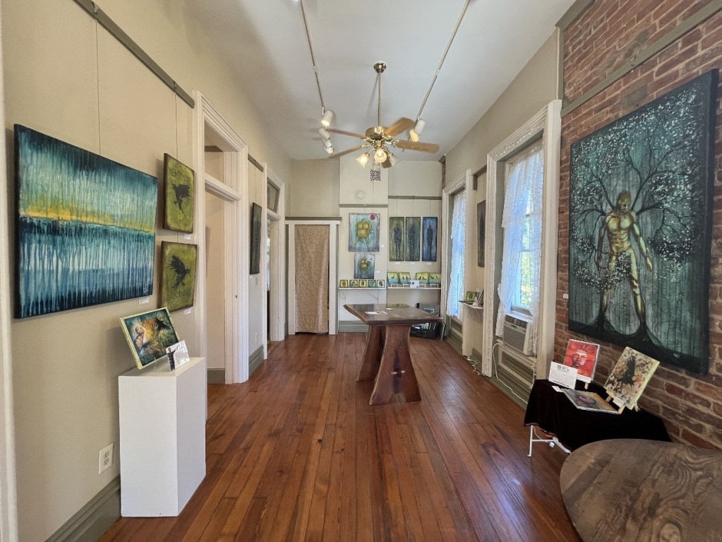 Interior view of an art gallery featuring vibrant paintings on brick walls, wooden floors, and natural light. The space includes a central table and various displays of artwork and promotional materials.
