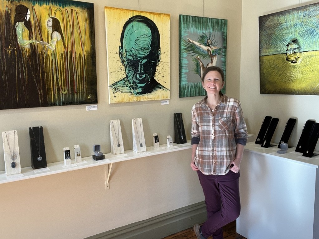 A woman stands in an art gallery, smiling beside vibrant paintings on the wall and displays of handcrafted jewelry on a shelf.