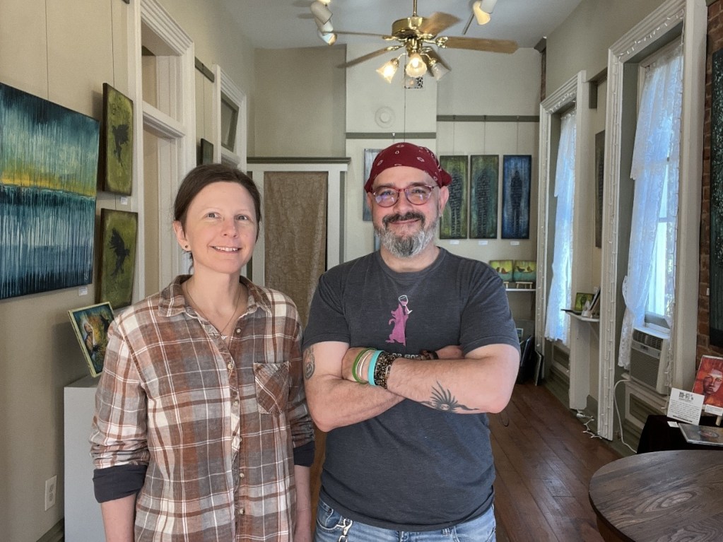 Two artists stand in a gallery space filled with colorful paintings on the walls. The atmosphere is inviting, featuring wooden floors and natural light from large windows.