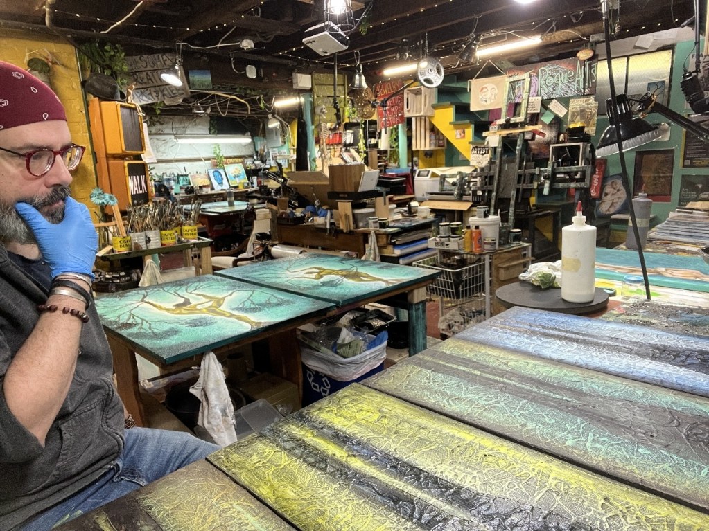 An artist wearing gloves and glasses is deep in thought at an art studio surrounded by various paintings and art supplies, with colorful walls and hanging lights creating a vibrant atmosphere.
