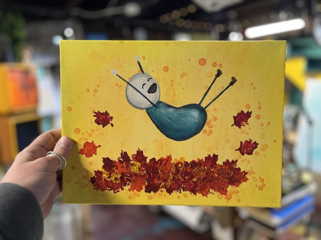 A person holding a whimsical painting of a character with a round blue body and a smiling face, surrounded by falling red and orange leaves against a bright yellow background.