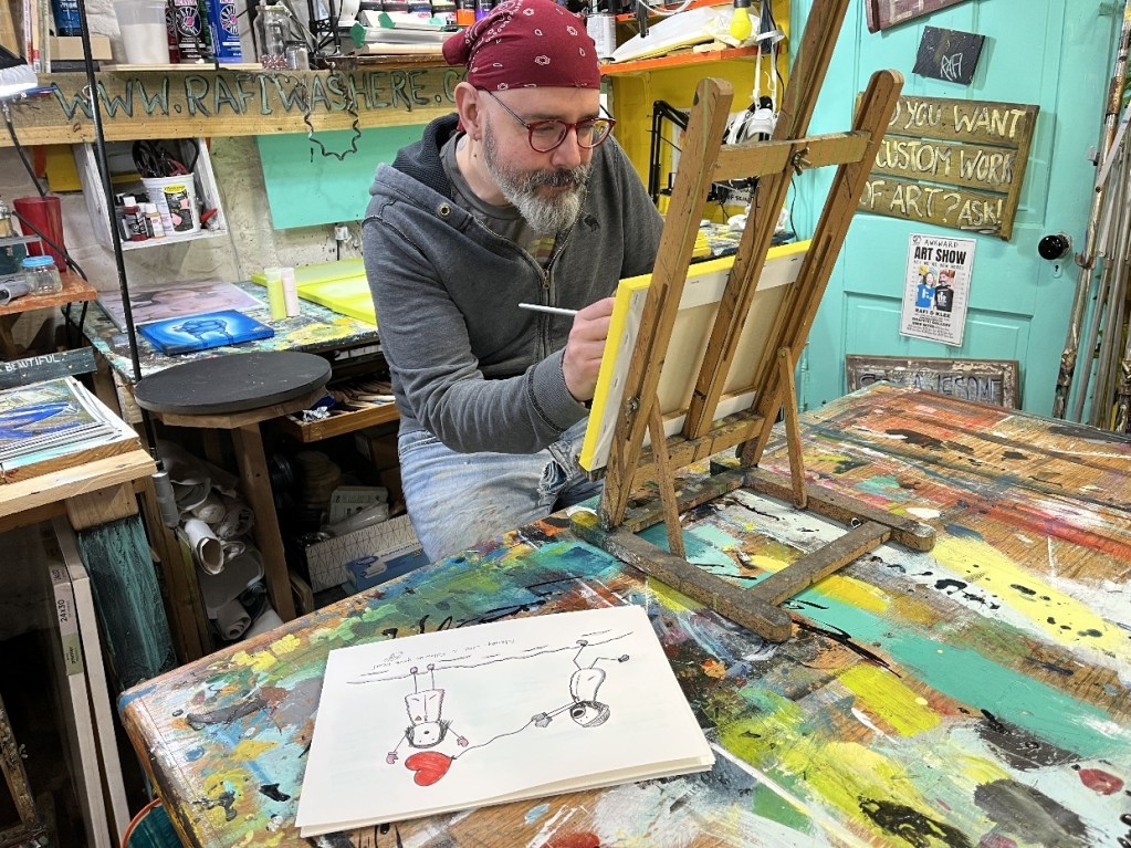 An artist sitting at a cluttered workbench, painting on a small canvas while sketching ideas on a nearby sheet of paper.