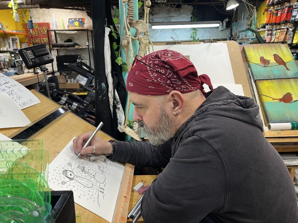 An artist wearing a bandana is sitting at a cluttered table, focused on drawing a whimsical illustration on paper. Various art supplies and colorful paintings are visible in the background, creating a creative atmosphere.