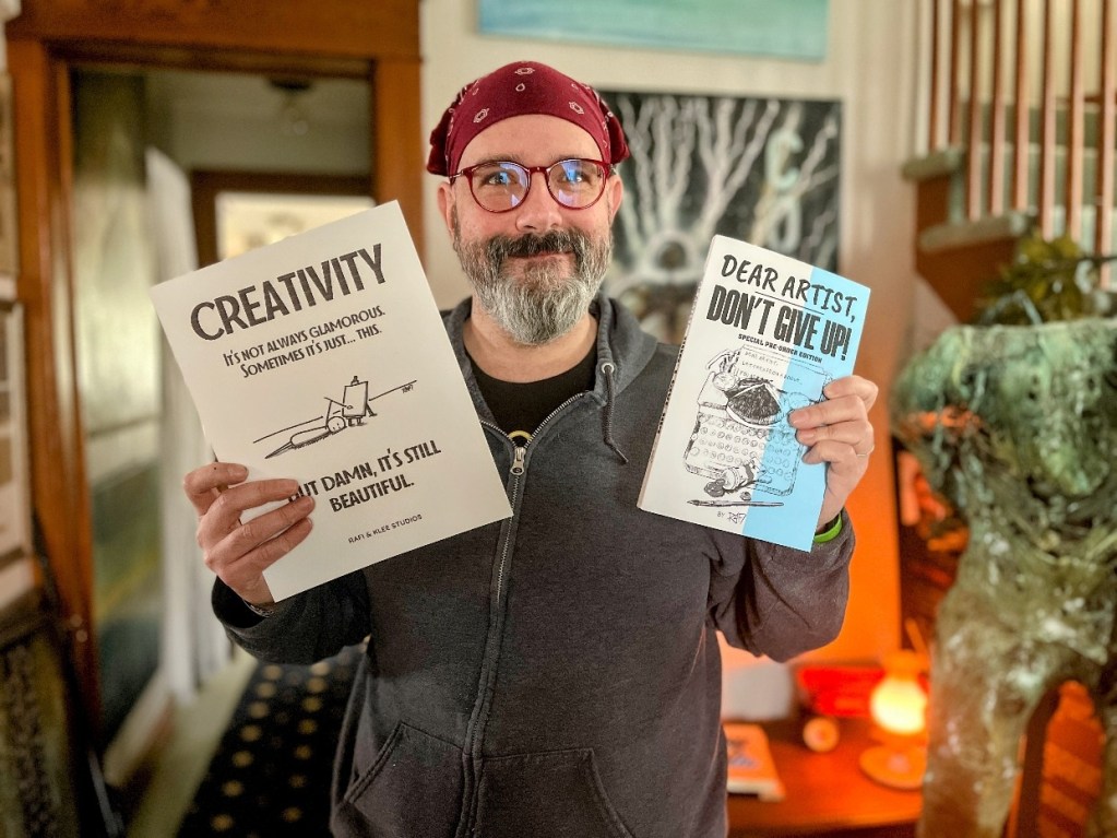 A person wearing a bandana and glasses, smiling while holding two books titled 'CREATIVITY: It's Not Always Glamorous' and 'DEAR ARTIST, DON'T GIVE UP!' in a cozy studio setting with artistic decor.