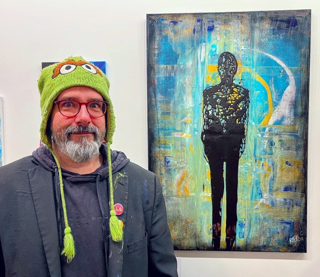 A person wearing a green hat with animal ears stands in front of a colorful abstract painting depicting a silhouetted figure.