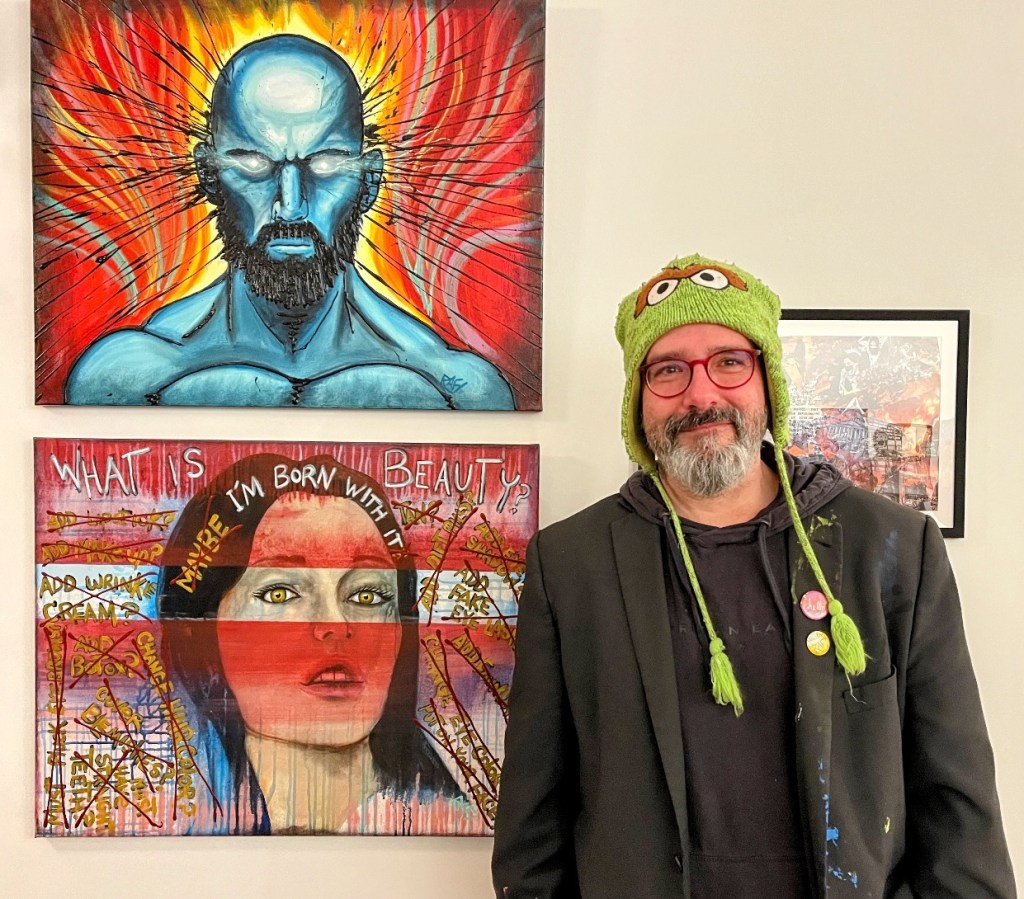 A man wearing a green hat and glasses stands in front of two colorful paintings, one depicting a blue figure and the other a woman with red and yellow elements, in an art exhibition setting.