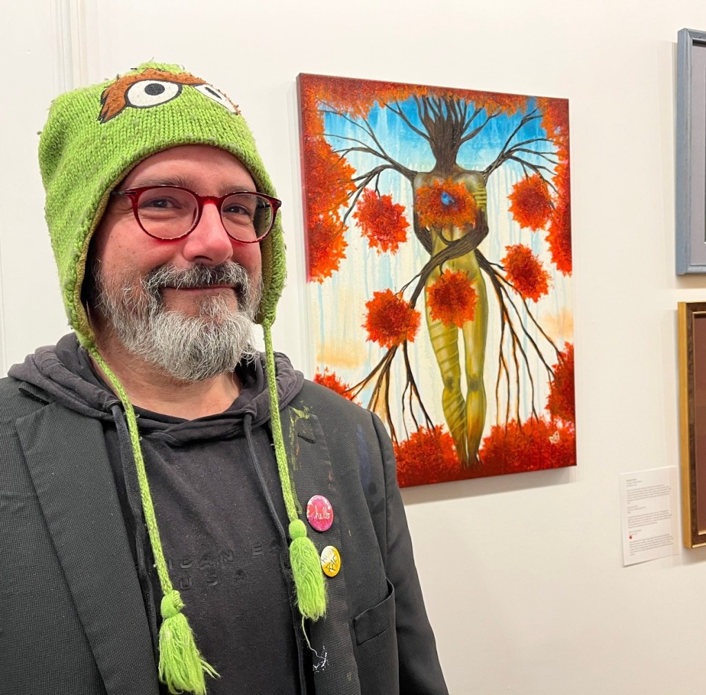 A person wearing a green beanie with eyes stands in front of an artistic painting of a figure with tree branches and colorful flowers.
