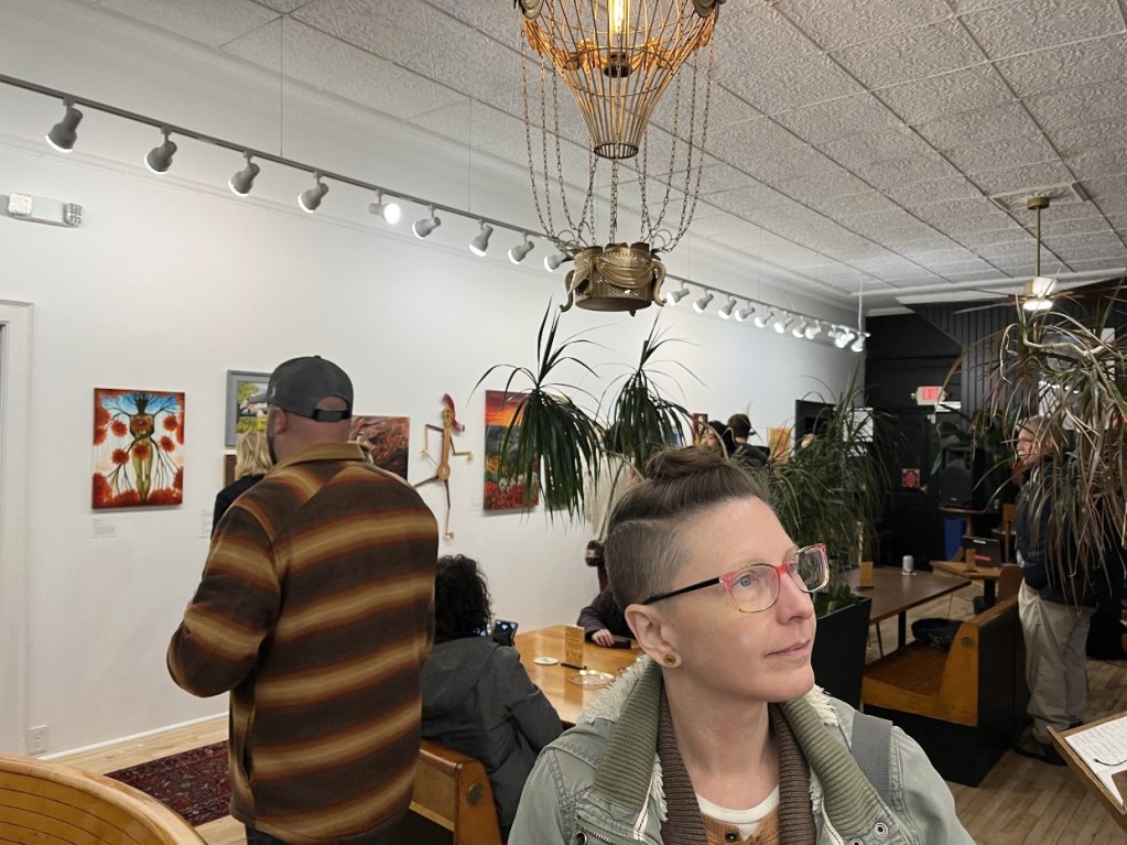 An art exhibition scene featuring attendees observing artwork on the walls, with a person in the foreground looking thoughtfully at the camera.