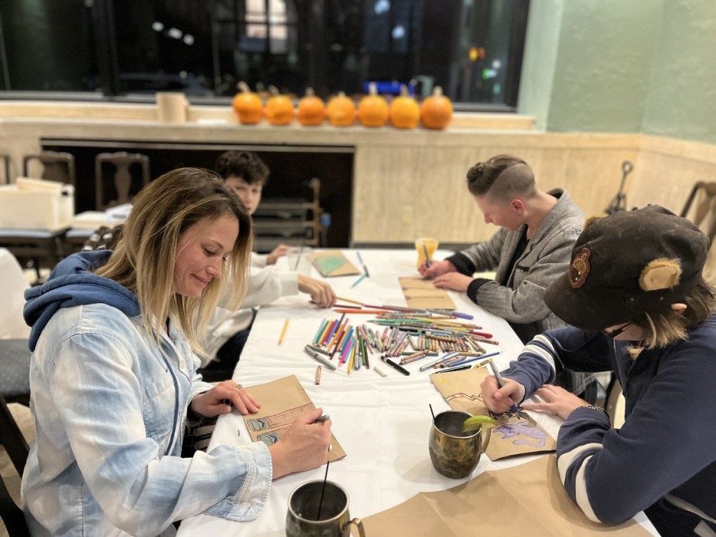 A group of four individuals engaged in a drawing activity at a table, surrounded by colorful art supplies. In the background, a row of pumpkins adds a festive touch to the scene.