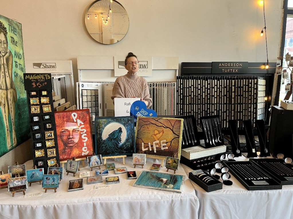 A vendor at an art pop-up event showcasing various art pieces and jewelry on a table, with colorful paintings and decorative magnets displayed prominently.