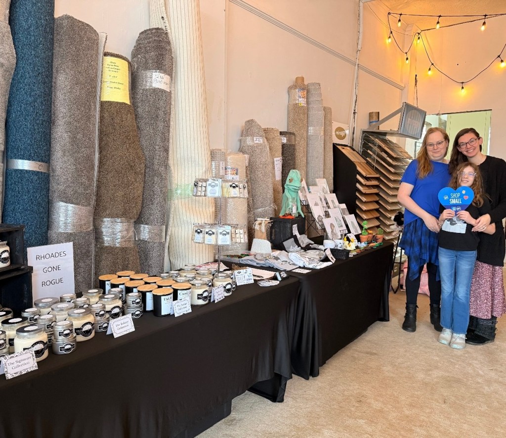 A group of three people, including a woman with glasses and a child, stand together in front of a display of various handmade items and candles at a pop-up shop. The backdrop features rolls of textured fabric and signs promoting small business shopping.