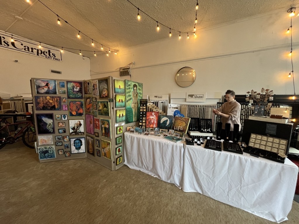 Artist pop-up event display featuring colorful artwork on panels and a jewelry table, with a vendor preparing in a brightly lit space decorated with string lights.