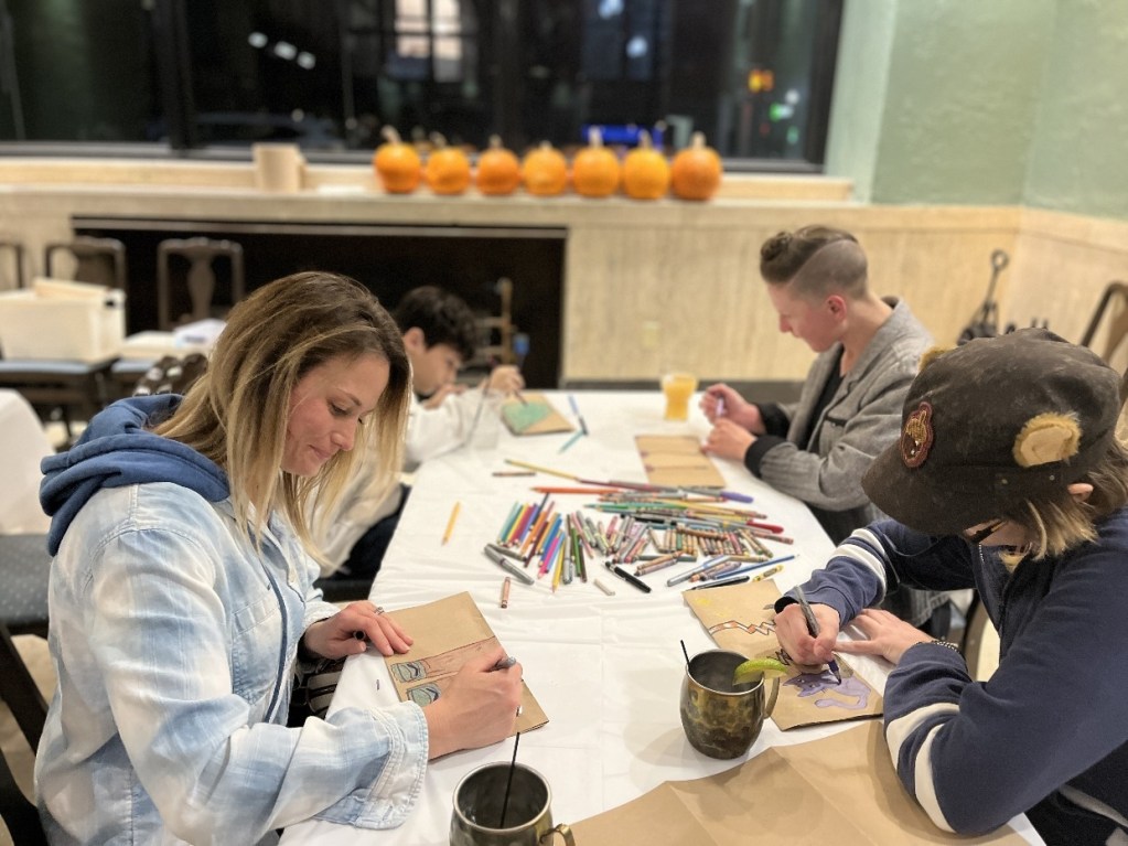 Four individuals engaged in a creative art session, drawing and coloring with various art supplies, including colored pencils, on a table filled with papers and drinks. Pumpkins are displayed in the background, adding a seasonal touch.