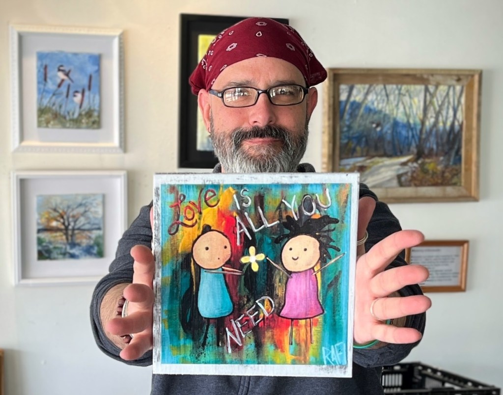 A person holding up a colorful painting that says 'Love is all you need,' featuring two whimsical figures. The background is vibrant and abstract, with several framed artworks displayed on the wall behind.