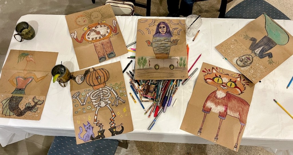 A table displaying colorful artwork featuring various imaginative characters, including a mermaid, a pumpkin, a skeleton, and a cat with large eyes, surrounded by art supplies.