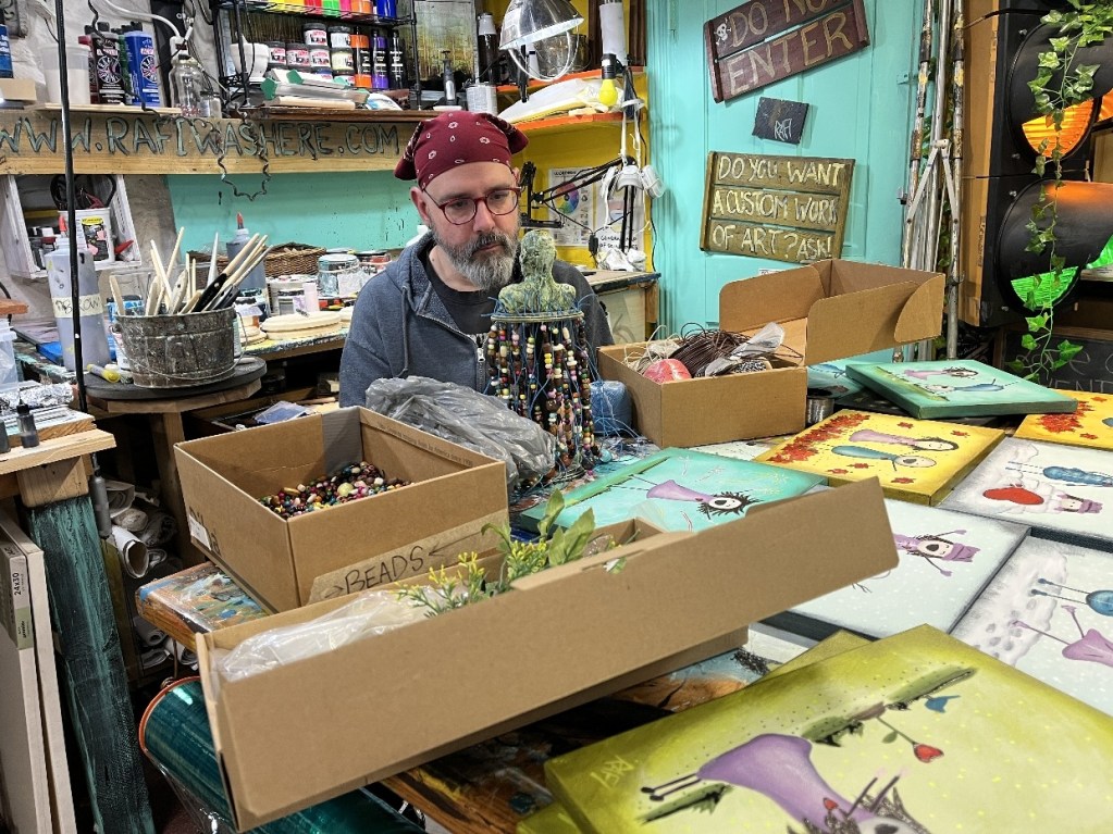 An artist sitting at a cluttered studio table surrounded by various art supplies, finished paintings, and open boxes labeled with craft materials.