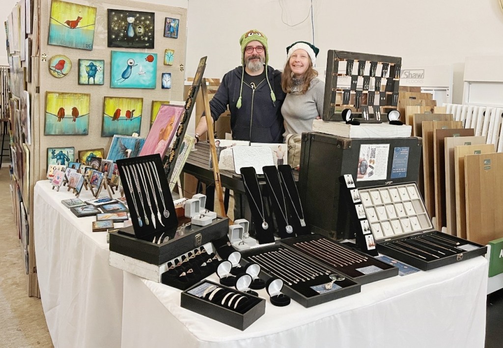 A smiling couple stands at their booth filled with handmade jewelry and colorful artwork at a craft fair.