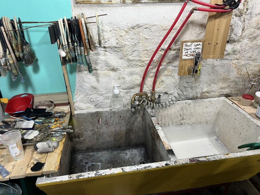 A cluttered artist studio sink area with various paintbrushes hanging on a rack, a utility sink showing signs of use, and a yellow painted base.