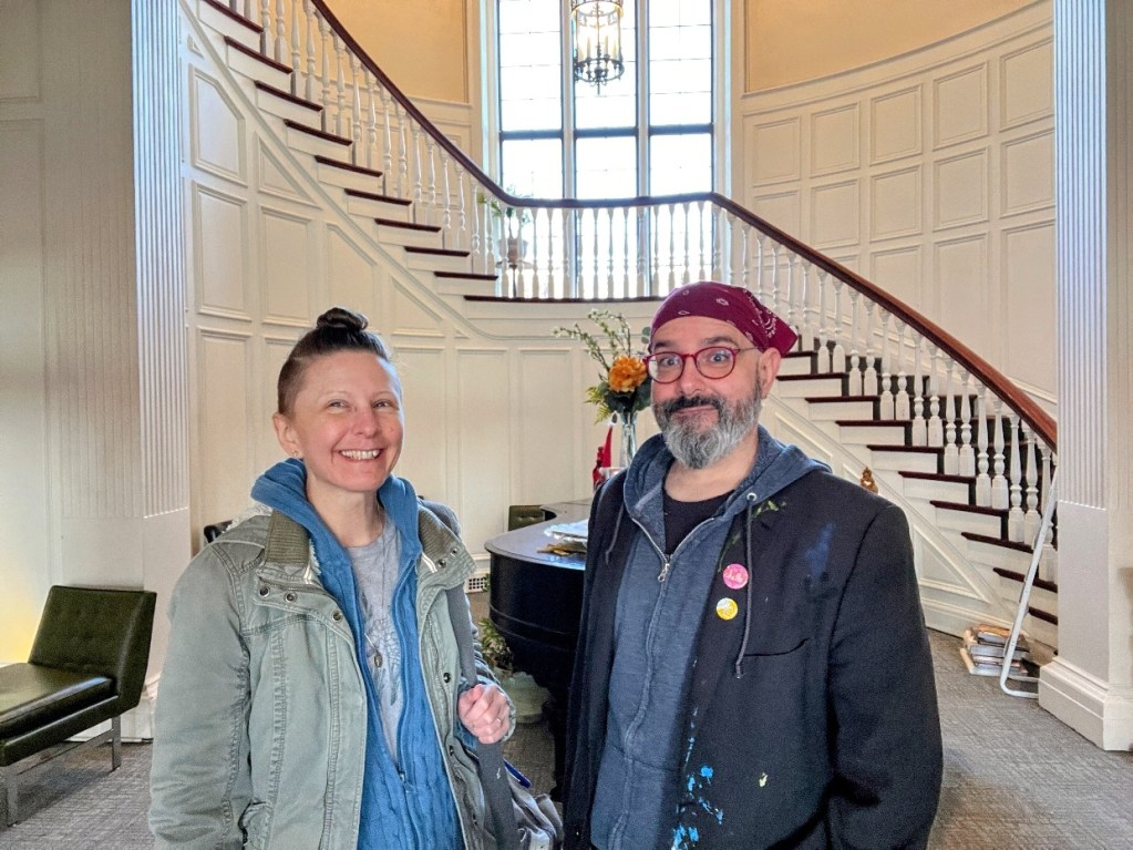 Two smiling individuals standing in a brightly lit room with a staircase in the background. One person has short hair and is wearing a casual jacket, while the other has a bearded face, glasses, and a bandana, wearing a t-shirt and a jacket with paint stains.